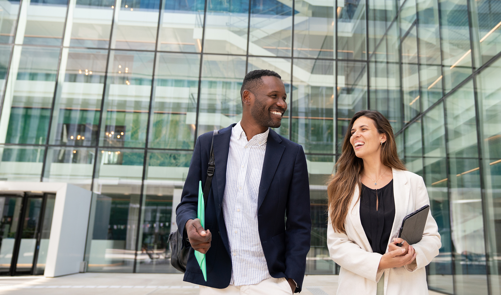 Two professionals, a man and a woman, are smiling and walking confidently in front of a modern glass office building. They represent the engaging opportunities and collaborative environment available in careers at Western Alliance.