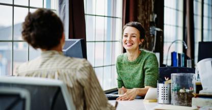Two business women sitting at a desk talking.
