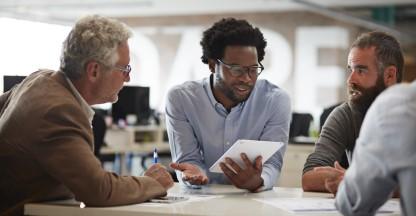 Black businessman holding a piece of paper and talking to two other men at a table. 