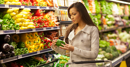 Woman in produce section of grocery store looking at a bell pepper.