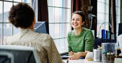 Two business women sitting at a desk talking.