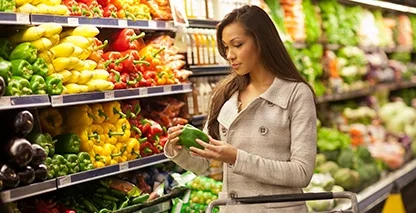 Woman in produce section of grocery store looking at a bell pepper.