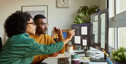 Two professionals collaborating at a desk, reviewing website designs and data on dual computer monitors.