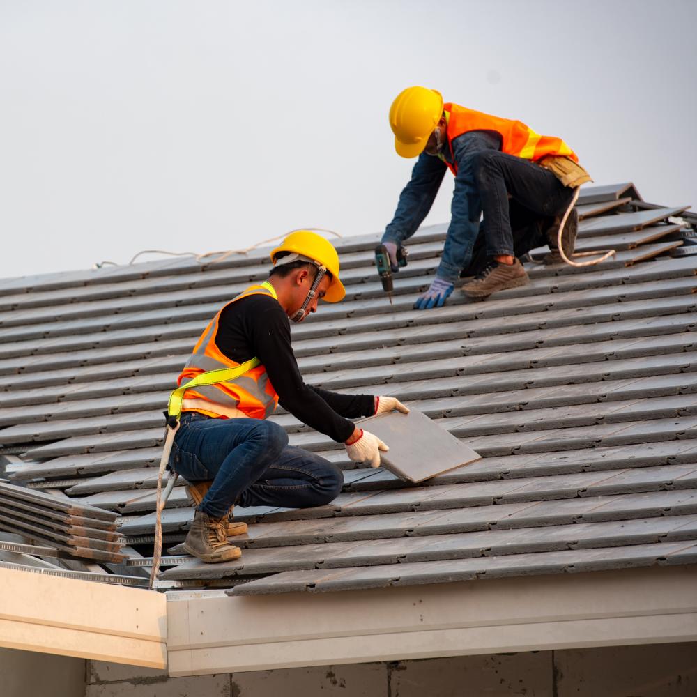 Two roofers in orange vests re-shingling a house roof.